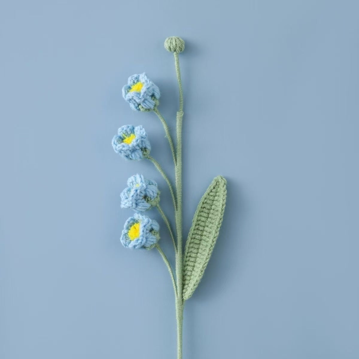 Crocheted Lily of the Valley puff flower branch with blue flowers and green leaves on a light blue background