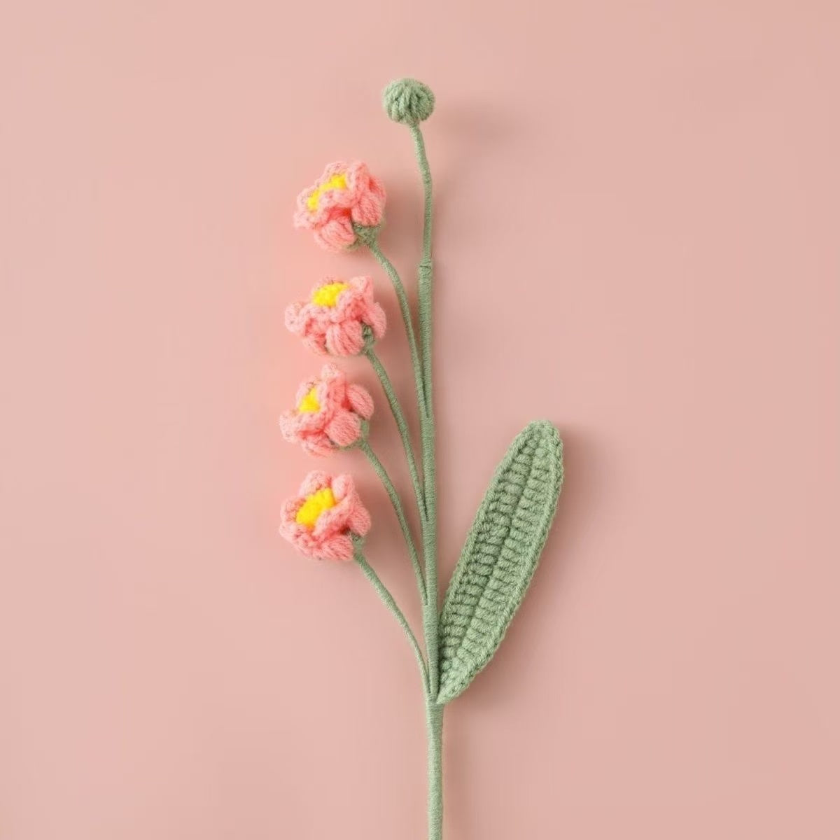 Crocheted Lily of the Valley puff flower with light pink petals and green leaves on a pink background