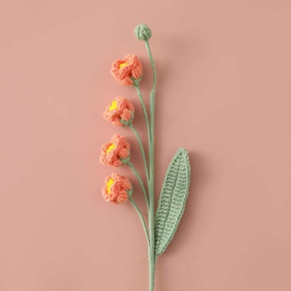 Knitted Lily of the Valley puff flower branch with orange flowers and a green leaf on a pink background